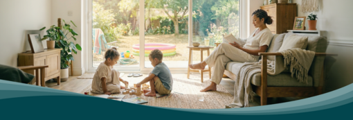 Zabraňte přehřívání interiéru díky vnitřnímu prostředí v rovnováze. Living room with large windows facing the garden. Children play on a rug while a person reads on the sofa. Zehnder residential ventilation ensures air exchange.